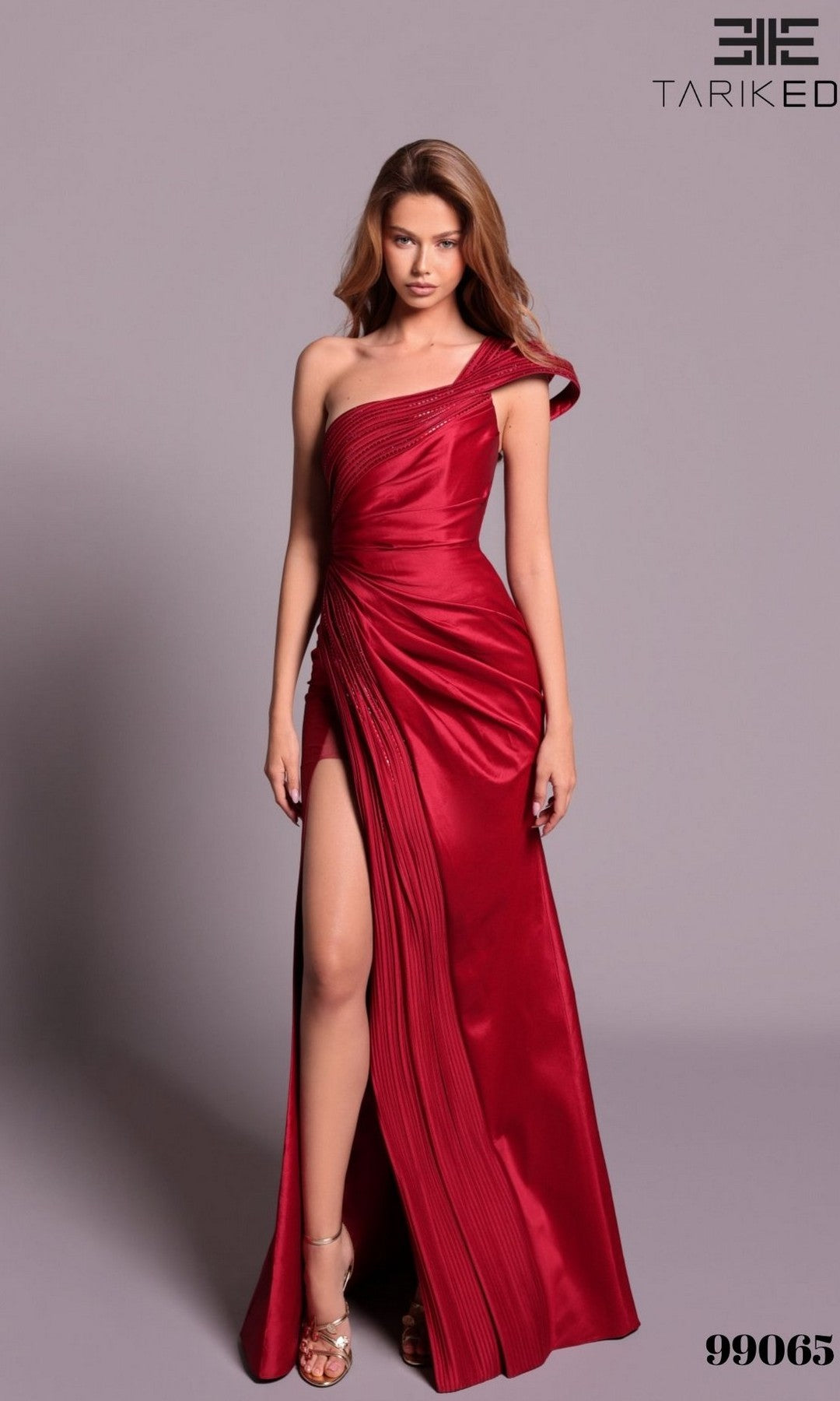 A woman models the Tarik Ediz 99065 long formal dress in burgundy, featuring a one-shoulder strap, pleated details, and a high leg slit, standing against a plain background.